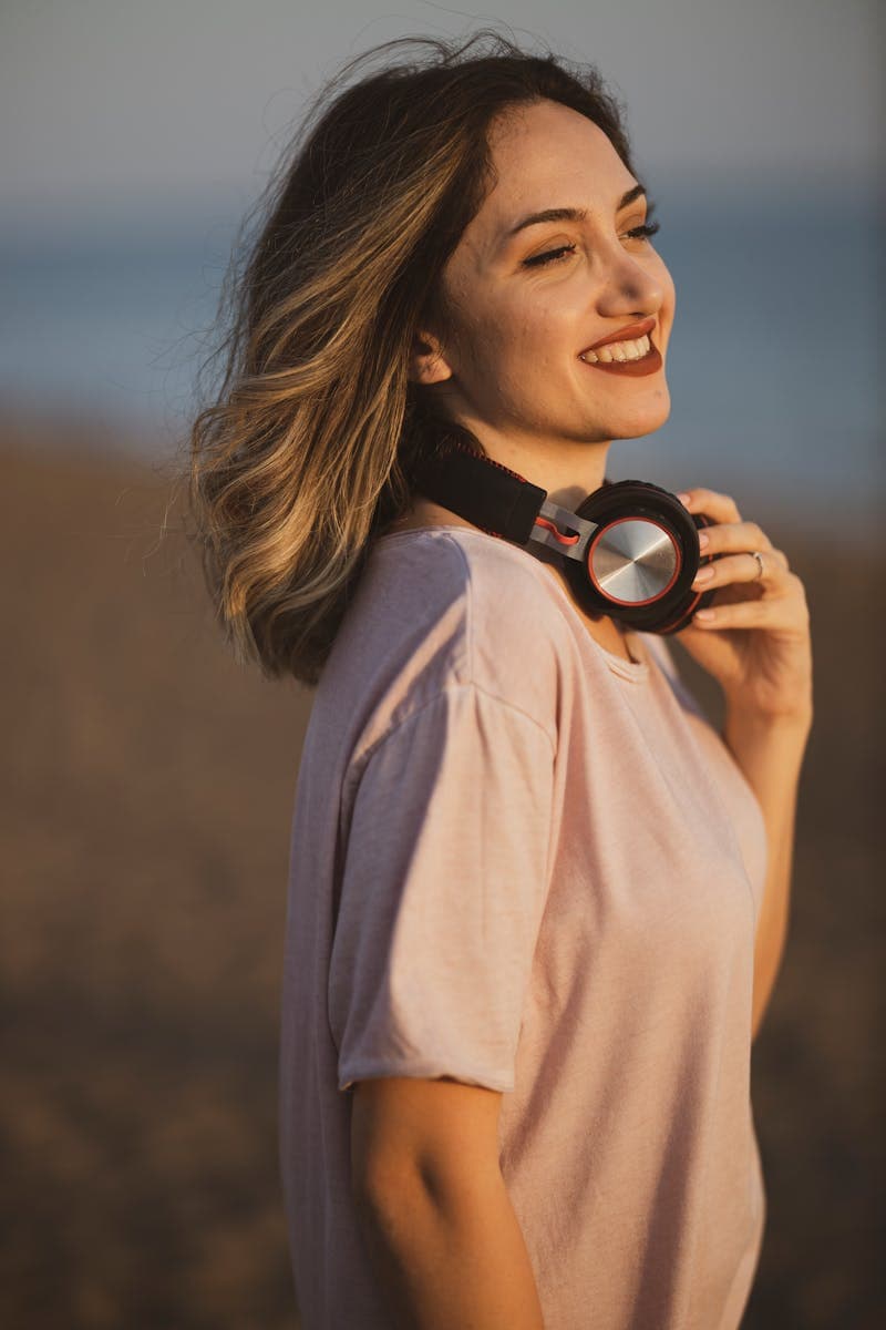 A woman enjoying outdoor music at sunset, showcasing stylish headphones and a casual pink shirt, perfect for outdoor leisure, summer, and active lifestyles.