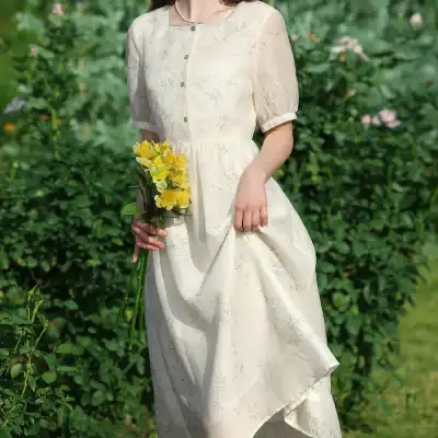 A woman wearing a white, embroidered dress with short sleeves, standing outdoors in a lush garden, holding a bouquet of yellow flowers, perfect for spring or summer occasions.