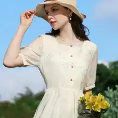 A woman wearing a light beige, embroidered summer dress with buttons, accessorized with a hat and jewelry, holding a bouquet of yellow flowers outdoors on a sunny day.