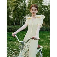 Bright, stylish woman in a yellow dress riding a bike outdoors surrounded by lush green trees, showcasing a fashionable look perfect for outdoor activities and lifestyle branding.