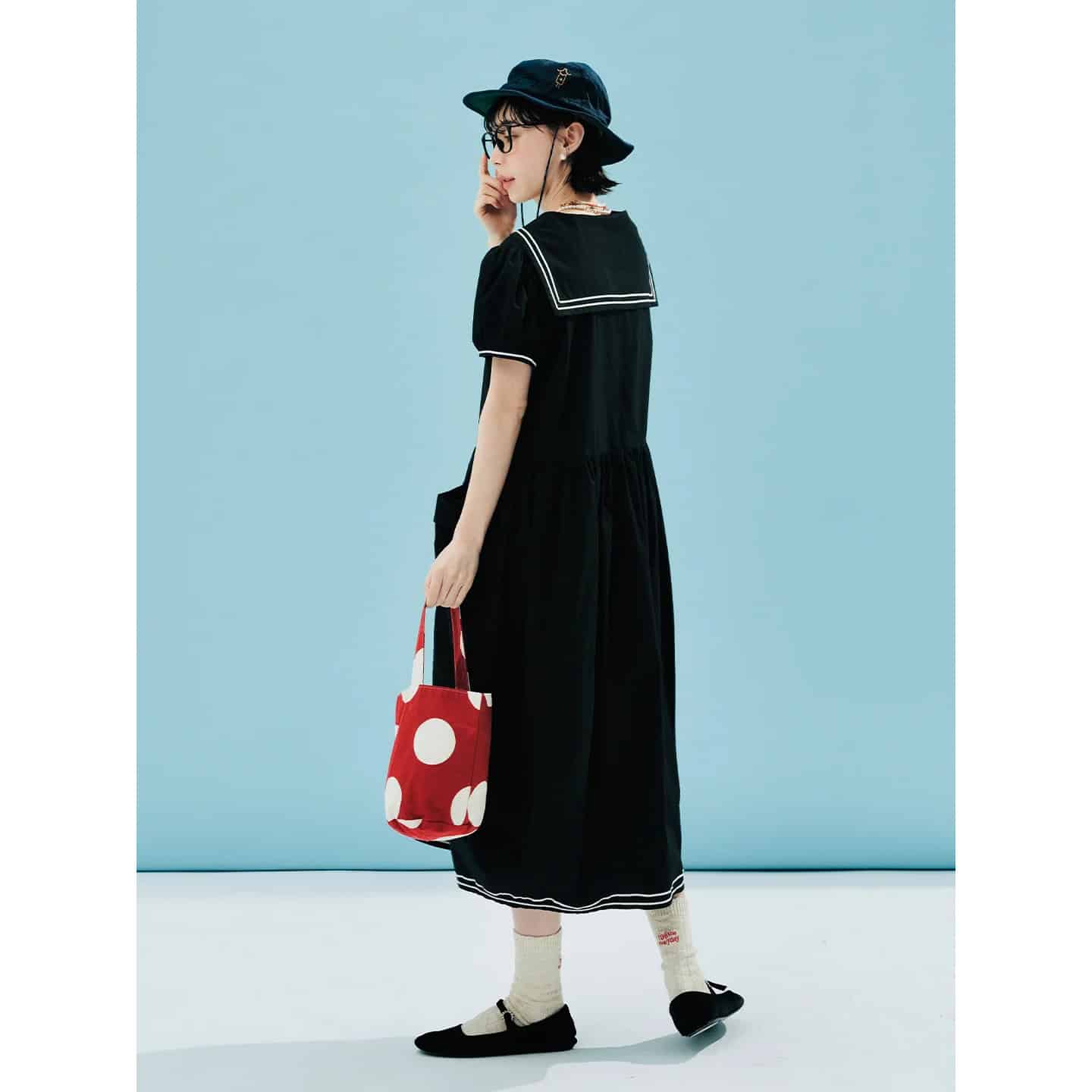 Stylish young woman wearing a black dress with sailor collar, black hat, glasses, and carrying a red polka dot tote bag, showcasing trendy accessories and apparel at the Monalisa medical fashion store.