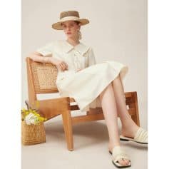 Stylish woman wearing a cream dress with a large collar, straw hat, and sandals, seated on a wooden bench; fashion, elegance, summer outfit for women.