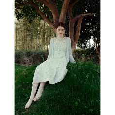 Young woman wearing a floral dress and light cardigan sitting on grass under a tree in a serene outdoor environment.