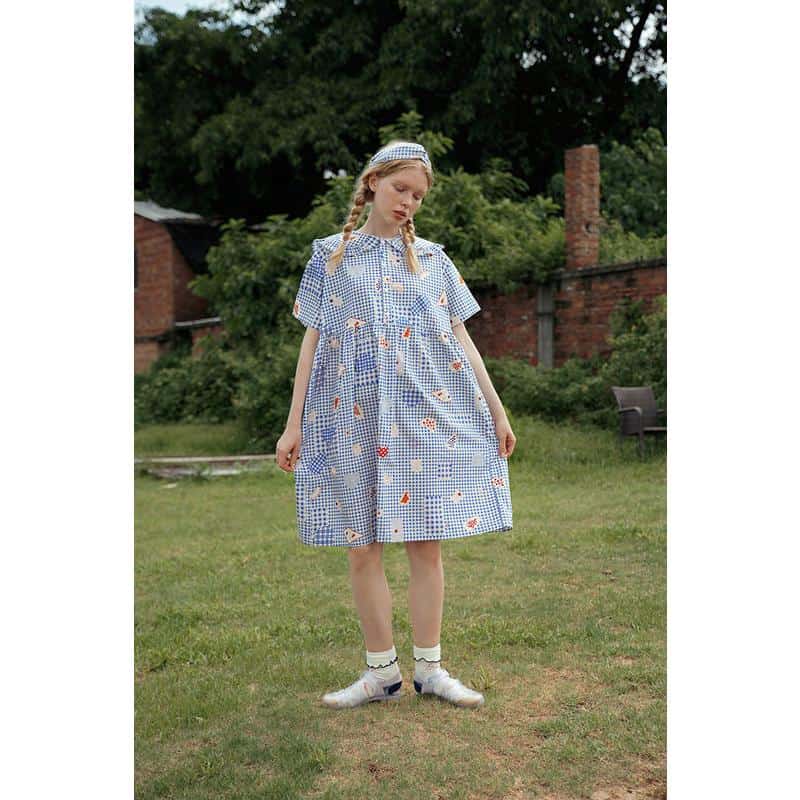 Young girl wearing a blue checkered dress with colorful patches, outdoor garden setting, casual fashion for kids, comfortable and stylish boys and girls clothing.