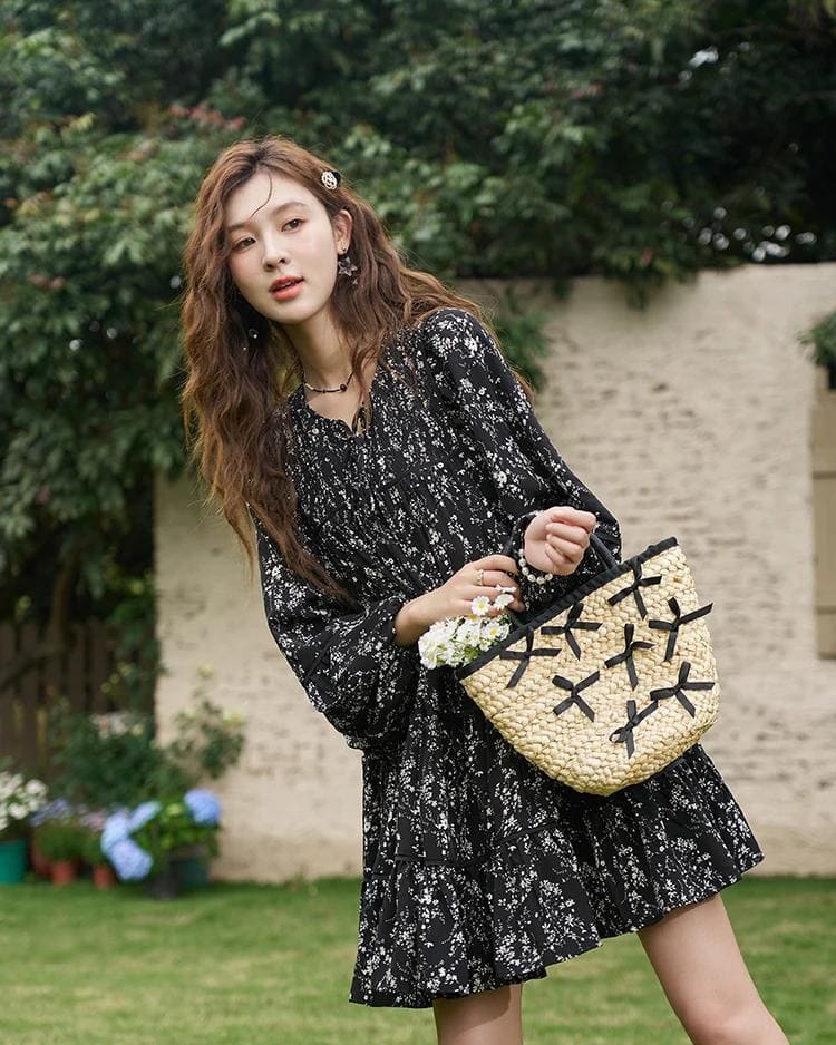 Stylish young woman holding woven handbag outdoors, wearing black floral dress, with lush garden background. Perfect for fashion, casual wear, and outdoor style inspiration.
