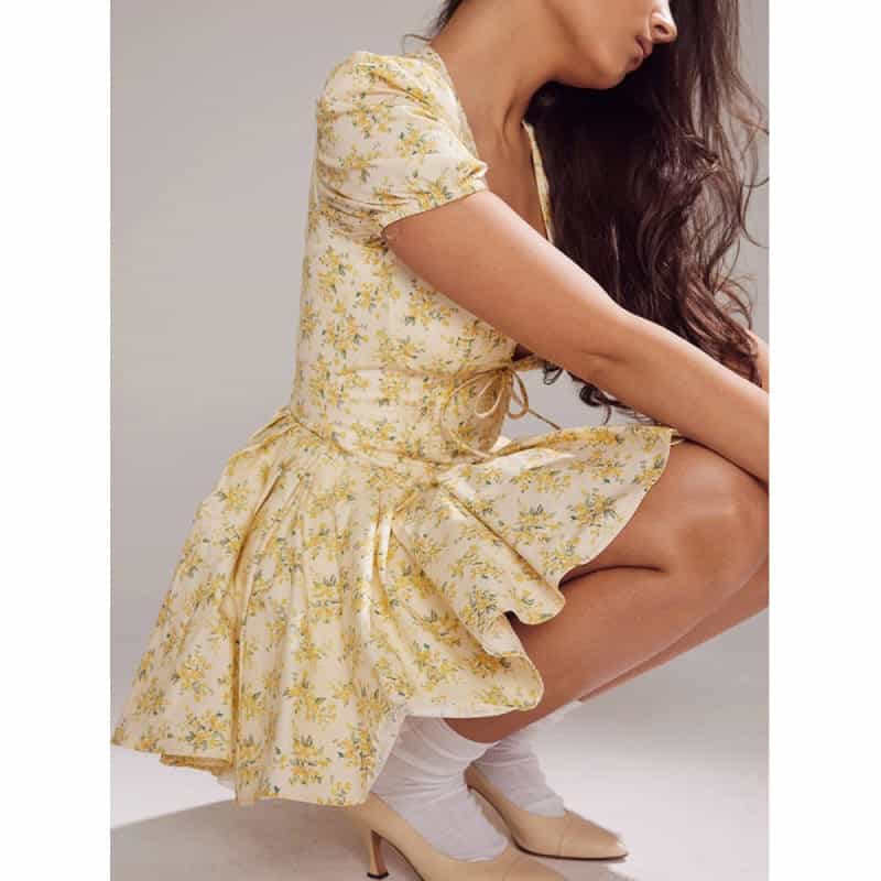 Close-up of a woman in a yellow floral dress, squatting with vintage heels and white socks. Elegant casual style with soft natural lighting and gray background.