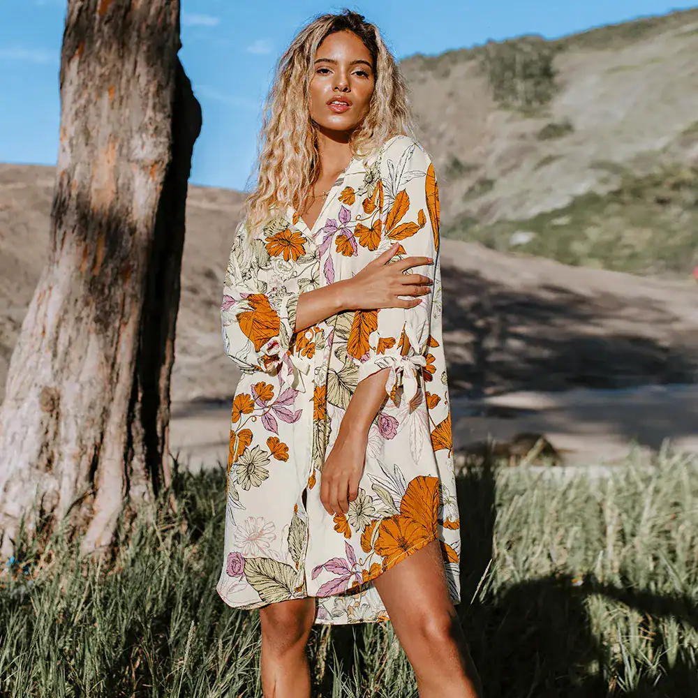 A stylish floral dress showcased outdoors, perfect for summer wear. Features vibrant orange, purple, and green floral patterns on lightweight fabric, ideal for a casual, chic look.