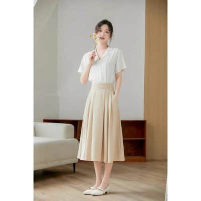 A stylish woman wearing a chic white blouse and beige pleated midi skirt, perfect for office or casual wear. Featuring comfortable flats and a minimalistic interior setting for versatile fashion.