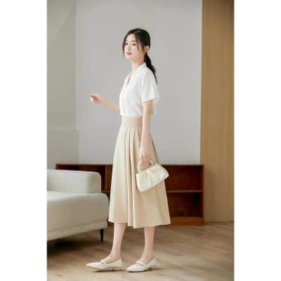 Image of a stylish woman dressed in a white blouse and beige pleated skirt, holding a chic purse, standing in a modern, minimalist healthcare or beauty clinic setting.