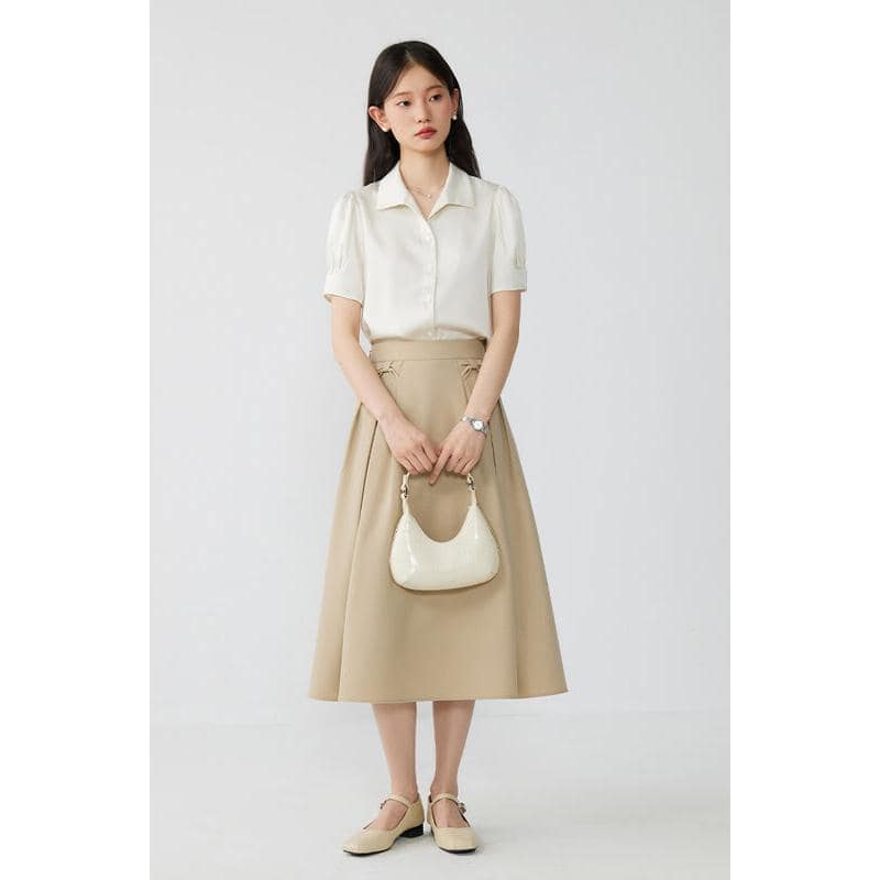 Elegant woman with beige skirt and white blouse holding a stylish cream handbag.