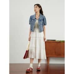 A stylish woman wearing a flowy white summer dress paired with a denim jacket, red sneakers, and carrying a striped tote bag, showcasing trendy fashion and sunglasses.