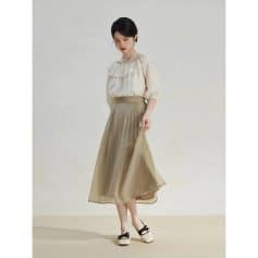A woman wearing a classy beige chiffon midi skirt paired with a ruffled cream blouse and stylish black-heeled shoes with bows, suitable for professional or formal settings.