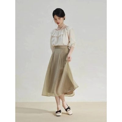 A woman wearing a classy beige chiffon midi skirt paired with a ruffled cream blouse and stylish black-heeled shoes with bows, suitable for professional or formal settings.