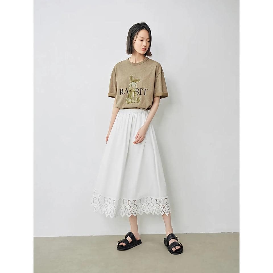 Stylish girl wearing a beige graphic T-shirt with "RABBIT" text and bunny illustration, paired with a white eyelet lace hem midi skirt and black sandals. Perfect for trendy, casual outfits.