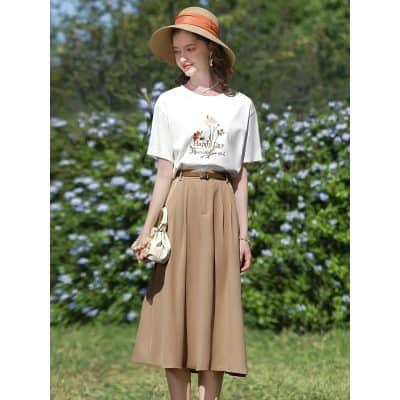 Stunning young woman wearing a stylish white T-shirt with floral embroidery and beige wide-leg skirt, accessorized with a sun hat and bag, outdoors in a lush garden setting.