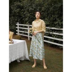 A woman in a stylish yellow blouse and floral skirt holding flowers outdoors at Monalisa Store. Perfect for spring fashion, casual wear, and elegant outdoor looks.
