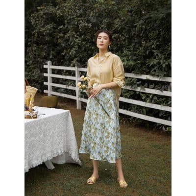 A woman in a stylish yellow blouse and floral skirt holding flowers outdoors at Monalisa Store. Perfect for spring fashion, casual wear, and elegant outdoor looks.