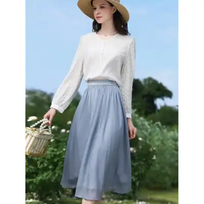 Cute woman in stylish white blouse and light blue flowy skirt, holding a woven basket against countryside background. Perfect for spring or summer fashion.