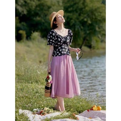 Beautiful woman enjoying sunny day by the river wearing stylish floral blouse and pink skirt, accessorized with sun hat and sunglasses, perfect for summer outings.