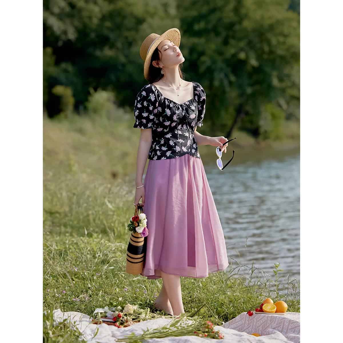 Beautiful woman enjoying sunny day by the river wearing stylish floral blouse and pink skirt, accessorized with sun hat and sunglasses, perfect for summer outings.
