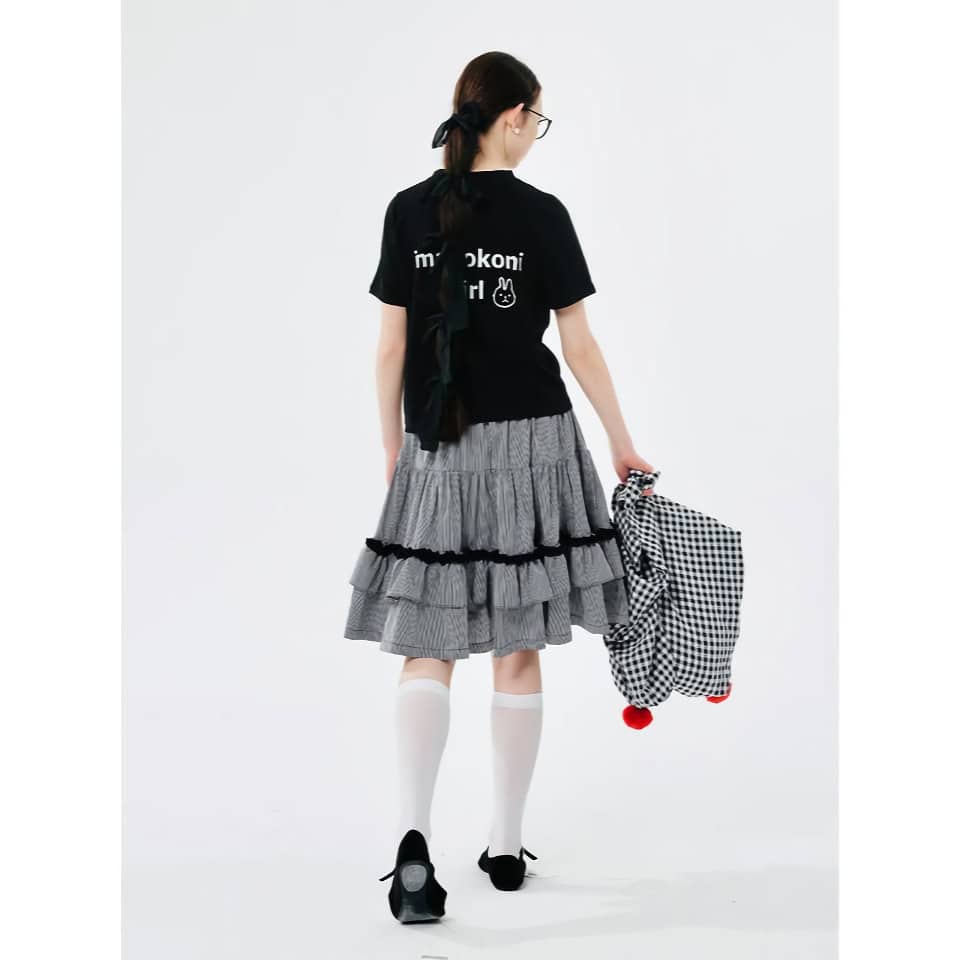 Young girl with long hair, glasses, and school bag, dressed in stylish skirt and black T-shirt, walking away.