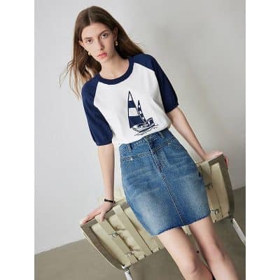A stylish woman in a nautical-themed white and navy blue top with a sailboat graphic, paired with a high-waisted denim skirt, sitting on a modern chair for a fashionable look.