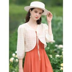 Beautiful woman wearing a white wide-brimmed hat, vintage-inspired white lace jacket, and coral-orange flowing dress; perfect for outdoor events, garden parties, or casual summer outings.