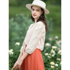 Young woman in stylish white lace blouse and coral skirt, wearing a wide-brimmed hat and pearl earrings, outdoor floral background, showcasing trendy women's fashion from Monalisa Store.