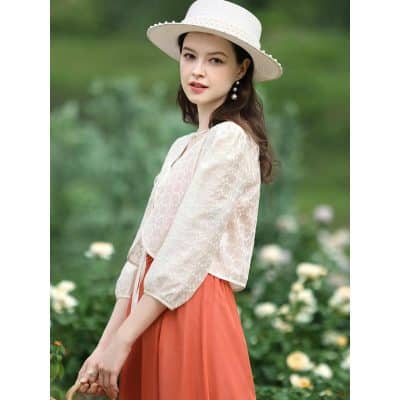 Young woman in stylish white lace blouse and coral skirt, wearing a wide-brimmed hat and pearl earrings, outdoor floral background, showcasing trendy women's fashion from Monalisa Store.