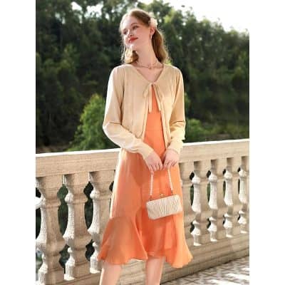 Young woman in stylish peach dress and cream cardigan, holding a chic beige purse with pearl strap, outdoors on a balcony - perfect for fashion and style lovers.