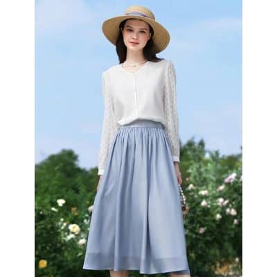 Stylish woman wearing a white long-sleeve blouse and blue maxi skirt, accessorized with a straw hat and pearl bag, standing in a garden with blooming flowers and blue sky in the background.