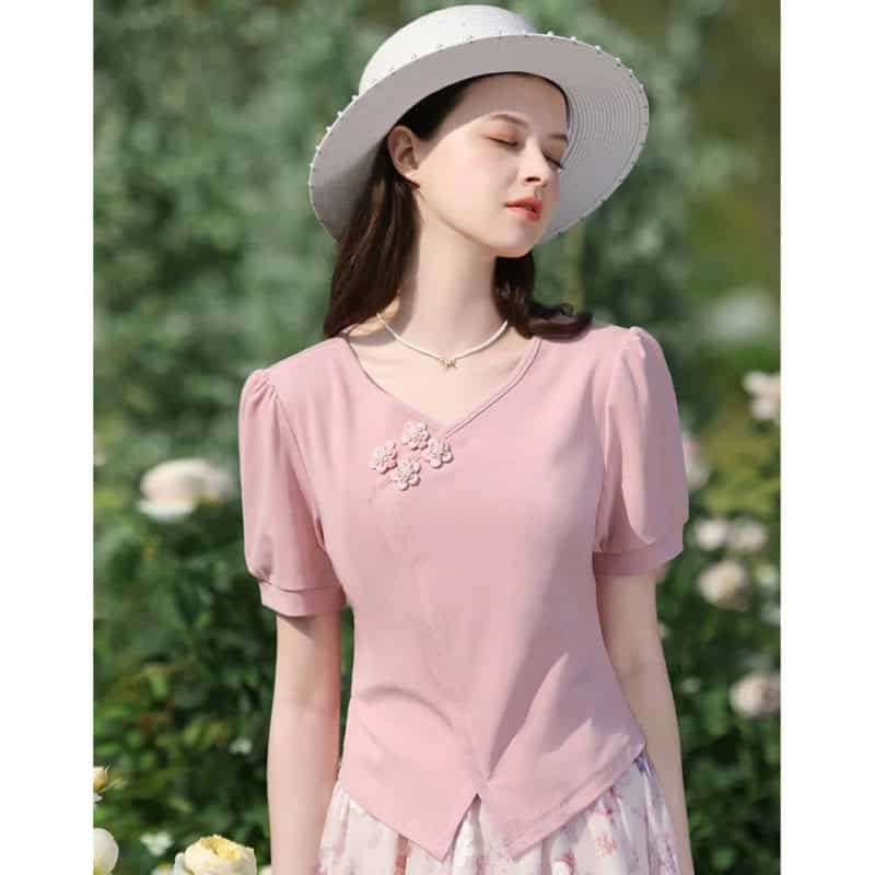 Young woman in pink blouse with embroidered flower detail, wide-brimmed hat, and outdoor garden setting, stylish and summer-ready.