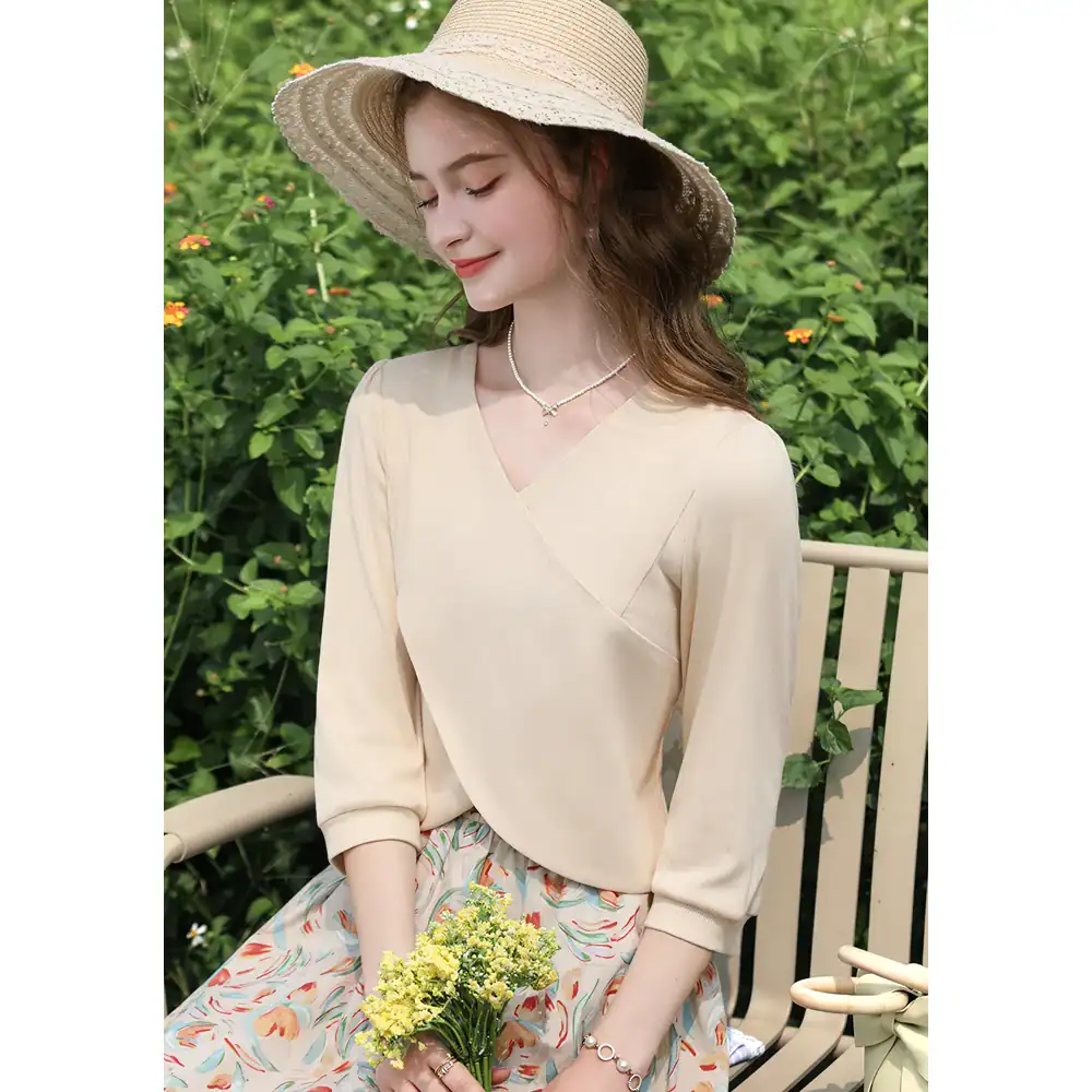 Stunning woman wearing a light beige summer dress with a flowy skirt, accessorized with a pearl necklace and a straw hat, sitting outdoors in a garden setting. Perfect for fashion and lifestyle bloggers.