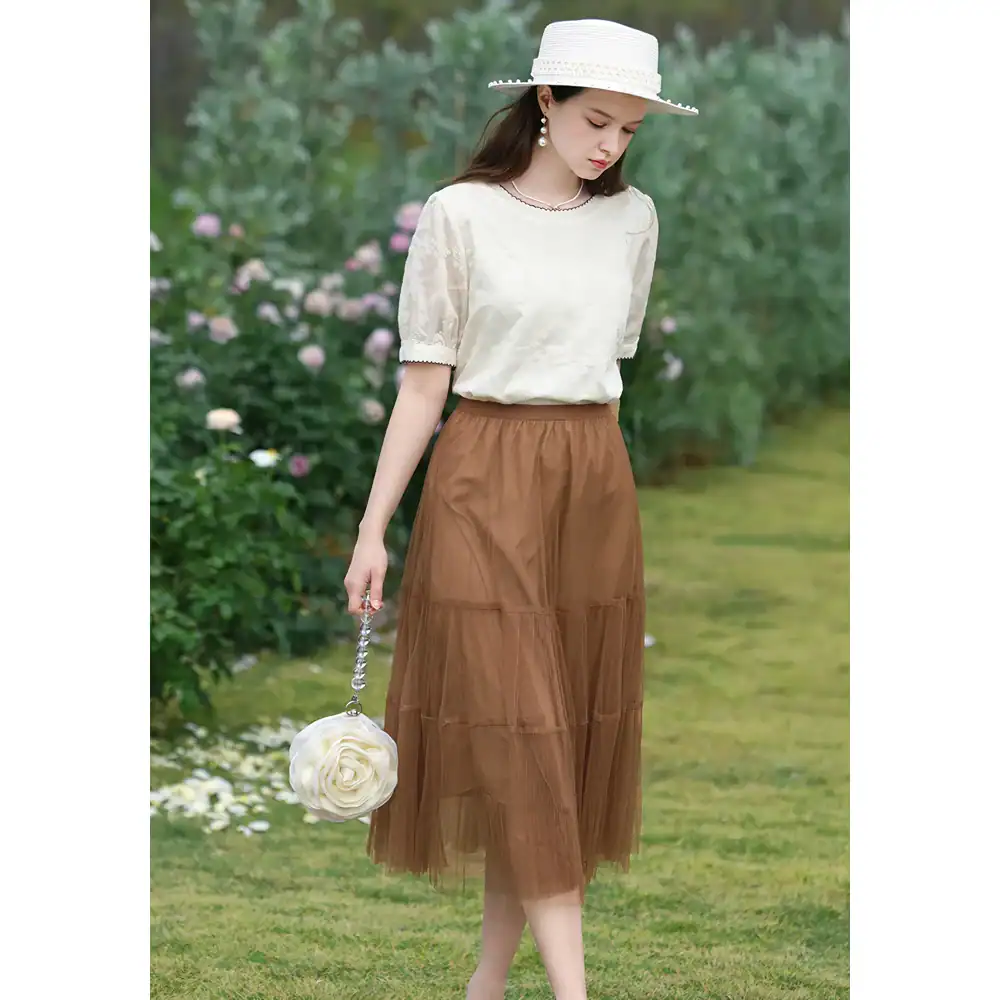 Stylish woman's summer outfit, flowy brown tulle skirt with cream blouse, wide-brim hat, and floral handbag, perfect for outdoor chic look and fashion-forward summer dressing.