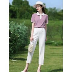 A fashionable woman wearing a pastel purple top, white trousers, and a chic hat. She holds a pearl handbag in a lush green outdoor setting, showcasing stylish summer fashion perfect for outdoor gatherings.