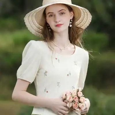 Beautiful young woman in a cream embroidered dress holding a bouquet of pink roses, wearing a wide-brimmed straw hat and pearl jewelry, outdoor garden setting.