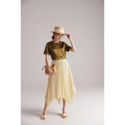 A stylish woman wearing a satin olive green top, flowy pleated cream-colored skirt, straw hat, and beige heels, showcasing chic outfit options from Monalisa Store in a minimal studio setting.