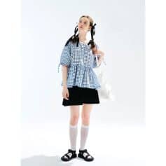 Cute young girl wearing fashionable gingham top, black skirt, knee-high socks, stylish sandals, and adorable hair accessories, posing in a clean, minimalist setting.