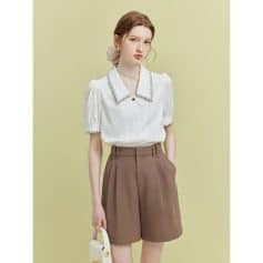 A stylish woman in a white blouse with detailed collar and brown high-waisted shorts against a soft green background. Perfect for office, casual, or chic outings.