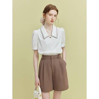 A stylish woman in a white blouse with detailed collar and brown high-waisted shorts against a soft green background. Perfect for office, casual, or chic outings.