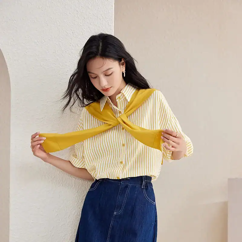 A stylish young woman wearing a yellow and white striped blouse with a yellow sweater draped over her shoulders, showcasing casual-chic fashion.