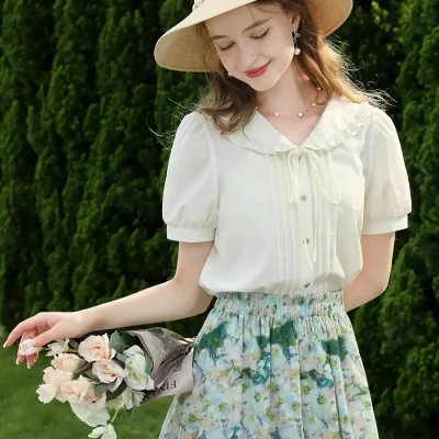Beautiful young woman in flowy floral skirt and white blouse, holding a bouquet of flowers, wearing a sunhat and jewelry, standing outdoors with lush greenery background.