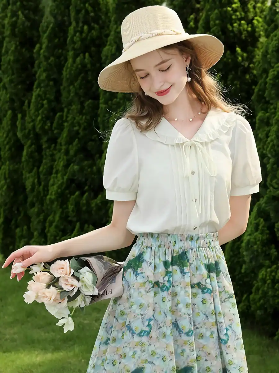 Beautiful young woman in flowy floral skirt and white blouse, holding a bouquet of flowers, wearing a sunhat and jewelry, standing outdoors with lush greenery background.