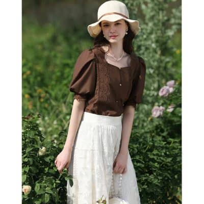 A stylish woman wearing a brown blouse with puff sleeves and lace details paired with a white flowing skirt, accessorized with a wide-brim hat and jewelry, standing outdoors in a lush garden setting.