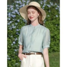 Stylish woman wearing hat and sunglasses, showcasing trendy summer fashion, light blue blouse, and outdoor greenery background. Perfect for summer sunglasses and fashion shopping needs.