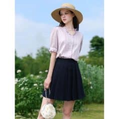 Stylish woman wearing a light pink blouse and a black pleated skirt, accessorized with a straw sun hat and floral handbag, outdoors in lush greenery. Perfect for summer fashion and casual outings.