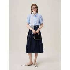 A fashionable woman in a light blue blouse and navy midi skirt, holding a black handbag, wearing glasses, stylish flats, ideal for professional and casual outings.