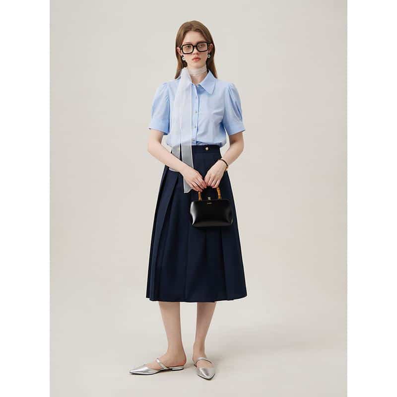 A fashionable woman in a light blue blouse and navy midi skirt, holding a black handbag, wearing glasses, stylish flats, ideal for professional and casual outings.
