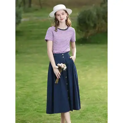 Young woman in a purple striped T-shirt, navy high-waisted skirt, and wide-brimmed hat, holding flowers outdoors, showcasing trendy fashion for women.