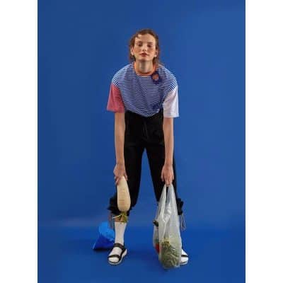 A young girl with wavy hair poses against a solid blue background, wearing a colorful striped t-shirt, black pants, and sneakers, holding a plastic bag with groceries and a water bottle, showcasing modern casual style.